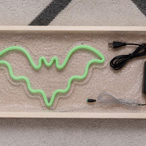 May include: A light green neon bat sign mounted on a clear acrylic backing, resting inside a rectangular wooden frame. The sign includes a black power adapter with a cord and a separate thin silver wire connector, all packaged for protection.