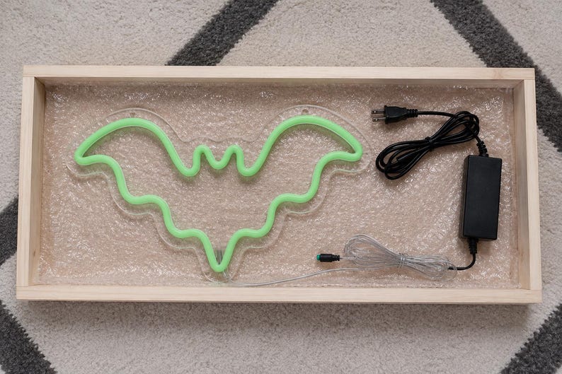 May include: A light green neon bat sign mounted on a clear acrylic backing, resting inside a rectangular wooden frame. The sign includes a black power adapter with a cord and a separate thin silver wire connector, all packaged for protection.