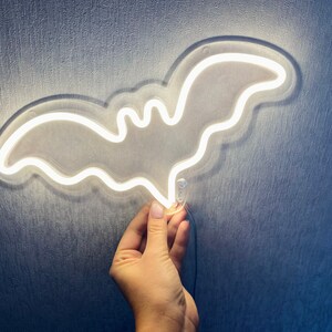 May include: A bat-shaped neon light fixture emitting a bright white glow. The light is constructed with a clear acrylic backing that follows the outline of the bat shape, and it includes a thin black power cord extending from the bottom edge.