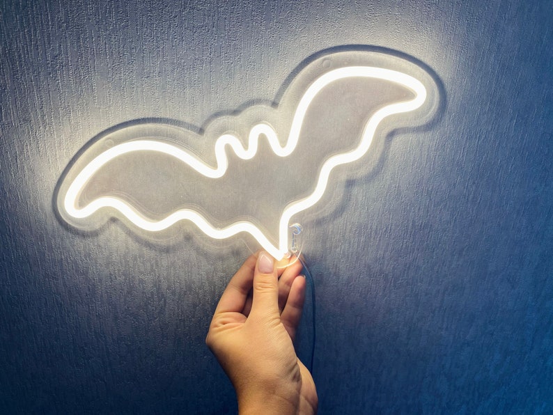 May include: A bat-shaped neon light fixture emitting a bright white glow. The light is constructed with a clear acrylic backing that follows the outline of the bat shape, and it includes a thin black power cord extending from the bottom edge.