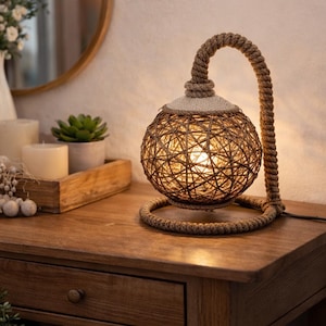 Peut inclure: Lampe de table décorative avec un abat-jour sphérique tissé et un support arqué enroulé de corde. La lampe est posée sur une surface en bois, et l'abat-jour est éclairé de l'intérieur. La base de la lampe est un design circulaire en corde.