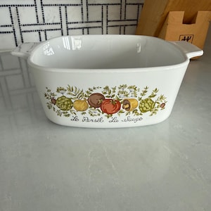 May include: White rectangular casserole dish with handles, decorated with a colorful vegetable and herb design. The words "Le Persil La Sauge" are written below the design.