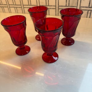 May include: Four ruby red vintage drinking glasses. Each glass has a flared shape with a detailed pattern and a rounded base. The glasses are arranged on a light-colored surface, showcasing their rich color and design.