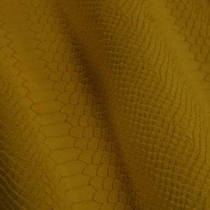 Op de afbeelding: Een close-up van een gele, textuurrijke stof met een krokodilleerpatroon.