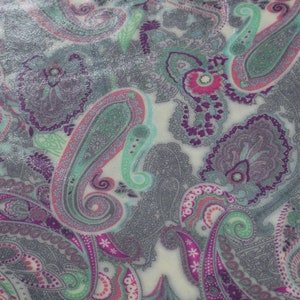 Può includere: Primo piano di un tessuto con un motivo paisley. Il design presenta grandi motivi a forma di goccia nei toni del grigio, viola, rosa e verde acqua, su uno sfondo color crema. Il tessuto ha una sottile lucentezza, che esalta i dettagli intricati.