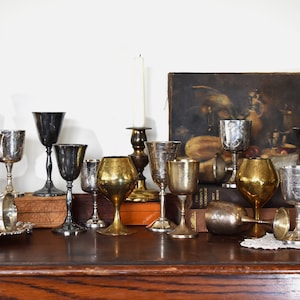 May include: A collection of antique goblets and chalices in various metallic finishes, including silver and gold. Some feature etched designs. A candle and a painting are in the background, all arranged on a wooden surface.