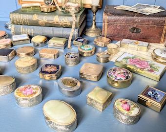 May include: A selection of vintage trinket boxes in diverse forms, such as round, oval, and rectangular. The boxes display elaborate designs, floral motifs, and metallic details. Some boxes are open, exposing their interiors. The items are arranged on a blue surface.