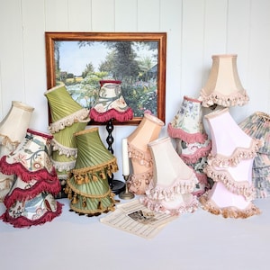 May include: A collection of various fabric lampshades featuring pleated and smooth textures in shades of green, pink, cream, and floral patterns. Many shades are adorned with decorative fringe or tassel trim along the bottom edges.