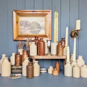 May include: A display of vintage stoneware bottles and jars in shades of brown and cream. A framed landscape painting, candles, and a brass candlestick holder are also present. The items are arranged on a wooden shelf and a blue surface.