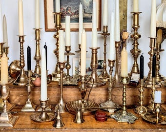 May include: A collection of various brass candlesticks of different heights and designs, each holding a taper candle in shades of white, cream, or black. The candlesticks are arranged on a wooden surface, featuring ornate bases and stems.