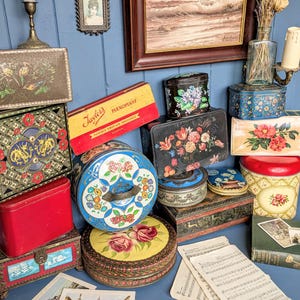 May include: A selection of antique metal tins in diverse forms and dimensions, adorned with floral, geometric, and landscape motifs. The tins showcase colours like red, blue, yellow, and black, with some bearing text. The display includes a "Taylors Flexoplast" tin.