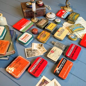 Op de afbeelding: Een verzameling vintage blikken en containers, waaronder EHBO-kits, keeltabletten en pastilles. De blikken zijn in verschillende kleuren, met tekstlabels en ontwerpen. Andere items zijn een kleine houten kist, een kandelaar en oude foto's.