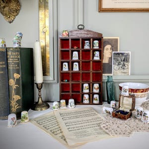 Puede incluir: Una vitrina de madera con 25 compartimentos llenos de dedalitos de porcelana en miniatura. Los dedalitos presentan diversos diseños, incluyendo patrones florales, animales y formas geométricas. La vitrina está sobre una mesa con partituras y otros dedalitos.