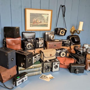 Puede incluir: Una colección de cámaras antiguas de varios tamaños y estilos, incluyendo cámaras de caja, telémetros y modelos compactos. Muchas cámaras tienen estuches de cuero. También se ven una impresión enmarcada y un candelabro.
