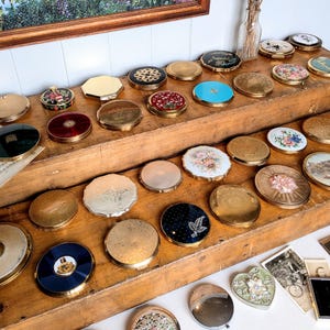 Puede incluir: Una colección de numerosos espejos de bolsillo vintage redondos y octogonales exhibidos en estantes de madera escalonados. Los polveras presentan diversos diseños que incluyen motivos florales, acabados metálicos y incrustaciones de esmalte.