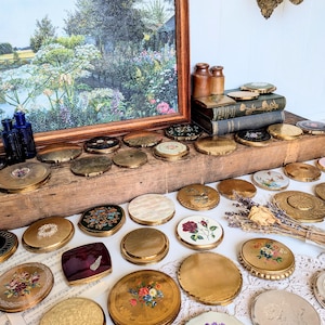 Op de afbeelding: Een grote collectie vintage ronde metalen poederdoosjes met diverse decoratieve deksels met bloemmotieven, gegraveerde texturen en emaille inleg, uitgestald naast twee kleine bruine flesjes en gestapelde antieke boeken.