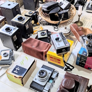 Puede incluir: Una colección de cámaras antiguas, incluyendo cámaras de caja y telémetros, en varios colores y diseños. Algunas cámaras están en sus cajas originales, y otras tienen estuches de cuero. La imagen incluye el texto "Kodak Instamatic 100" y "Coll 44".
