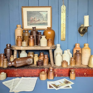May include: A display of antique stoneware jars and bottles in shades of brown and cream. The collection is arranged on a wooden stool and a red shelf. A framed print, sheet music, a brass wall sconce, and a decorative hanging piece are also visible in the background.