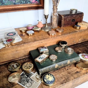 Puede incluir: Una colección de polveras de metal vintage y pequeñas cajas de baratijas con varias tapas decorativas con diseños florales, escénicos y de camafeo, dispuestas sobre una superficie de madera junto a un libro verde y fotografías en blanco y negro.