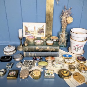 May include: A display of antique trinket boxes in diverse shapes and sizes, including round, oval, and rectangular. The boxes showcase intricate designs, floral patterns, and metallic accents. A vintage postcard and a brass ruler are also visible.