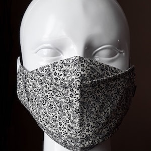 May include: A white face mask with a black and white paisley pattern. The mask is made of fabric and has a pleated design.