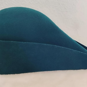 以下が含まれることがあります： 独特な曲線デザインのティール色の帽子。丸みを帯びたクラウンと、幅広で下向きに傾斜したつばが特徴です。柔らかいフェルトのような素材で作られています。ヴィンテージまたはレトロなスタイルである可能性があります。