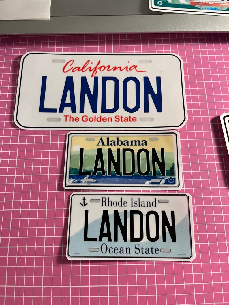 May include: Three license plate stickers with the name "LANDON". The top sticker is from California, the middle from Alabama, and the bottom from Rhode Island. Each plate has a different background and design.