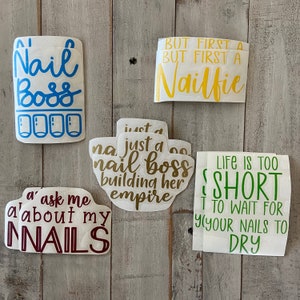 Può includere: Una collezione di adesivi in vinile bianco con varie frasi relative alla cura delle unghie. Gli adesivi presentano testo in blu, giallo, verde e bordeaux. Le frasi includono "Nail Boss", "But first a Nailfie" e "Life is too short to wait for your nails to dry."