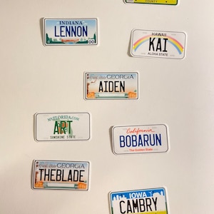May include: A collection of license plate-shaped stickers with various names and text. The stickers feature designs from states like Iowa, California, Hawaii, and Georgia. Some include names like "LENNON", "AIDEN", and "CAMBRY".