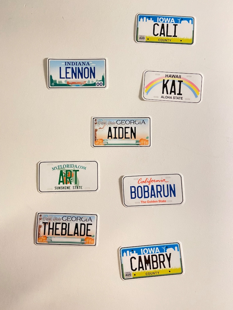 May include: A collection of license plate-shaped stickers with various names and text. The stickers feature designs from states like Iowa, California, Hawaii, and Georgia. Some include names like "LENNON", "AIDEN", and "CAMBRY".