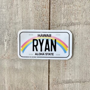 May include: A white license plate-shaped sticker with rounded corners. The word "HAWAII" is at the top, with "RYAN" in bold black letters below a rainbow arc. "ALOHA STATE" is at the bottom. The sticker is on a light wood-grain surface.