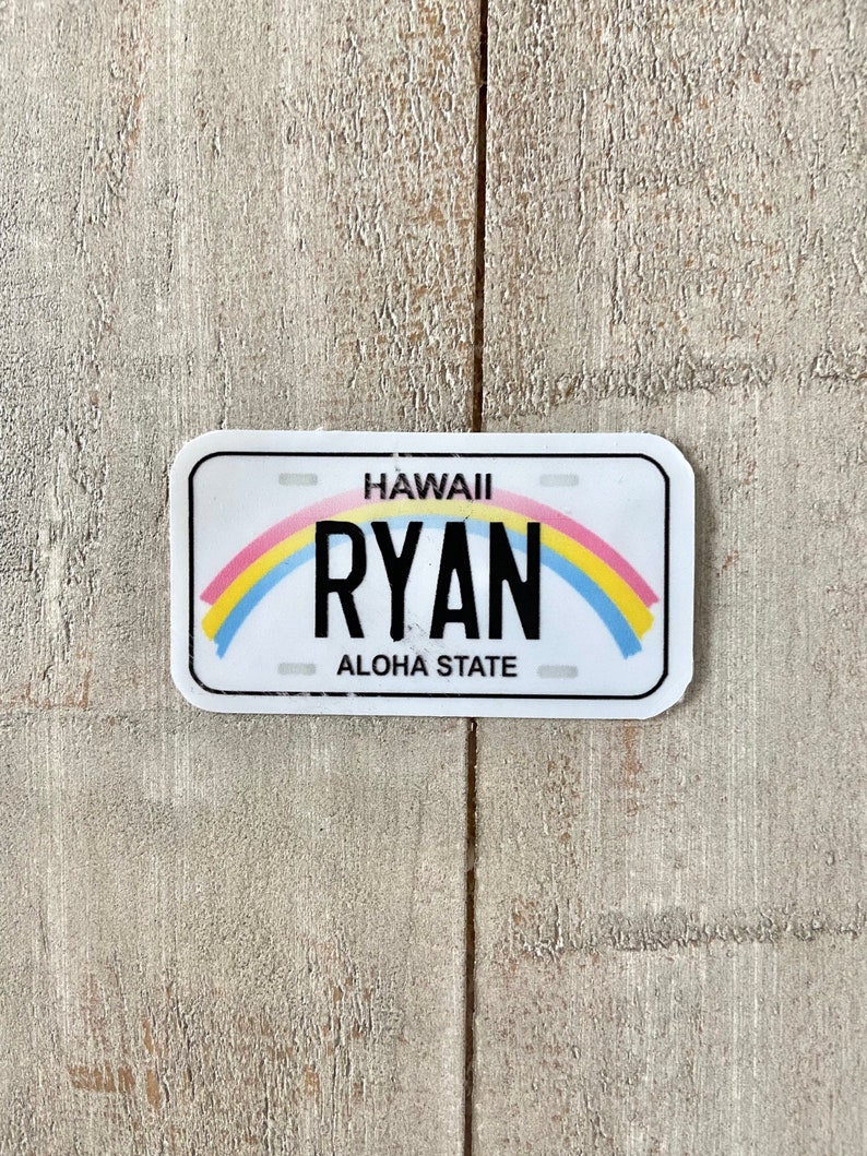 May include: A white license plate-shaped sticker with rounded corners. The word "HAWAII" is at the top, with "RYAN" in bold black letters below a rainbow arc. "ALOHA STATE" is at the bottom. The sticker is on a light wood-grain surface.