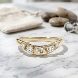 May include: A gold ring with a curved band, set with multiple clear gemstones of varying shapes. The gemstones include round, pear, and baguette cuts. A small beige pouch and a magnifying glass are in the background.