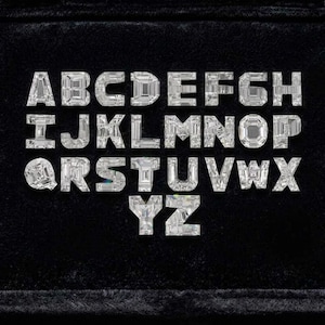 May include: A full alphabet of sparkling, clear gemstone letters, each with a unique faceted design, arranged on a black velvet surface. The letters are evenly spaced and presented in a neat, organized display.