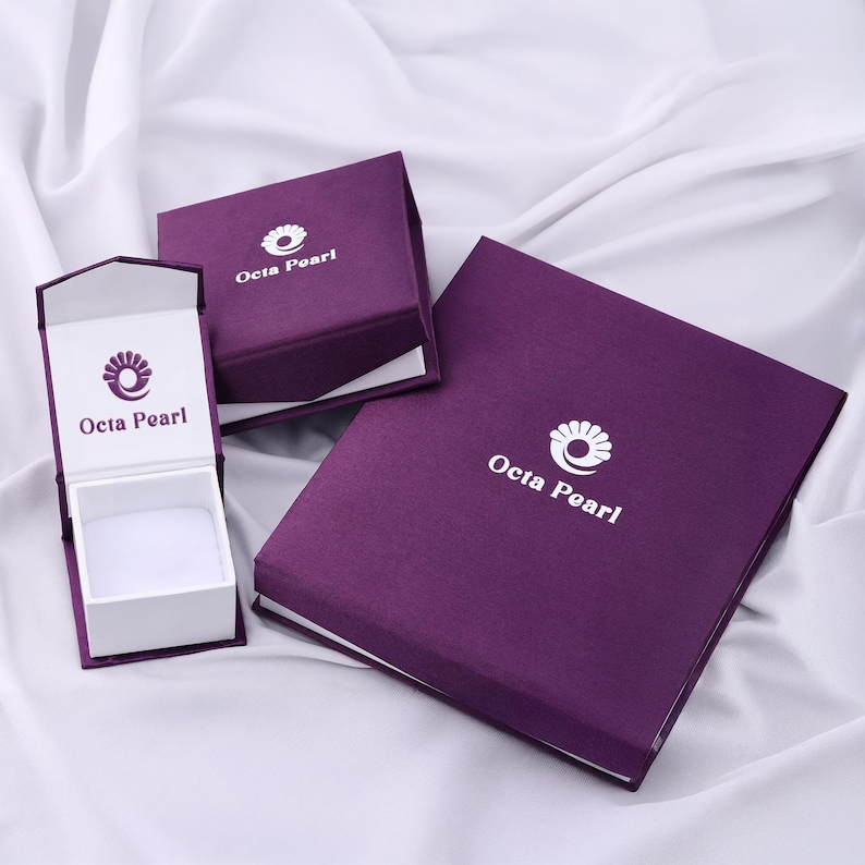 May include: An open purple jewelry box with a white interior sits next to two closed purple boxes. The boxes have the text "Octa Pearl" and a logo printed in white. The boxes are on a white, textured surface.