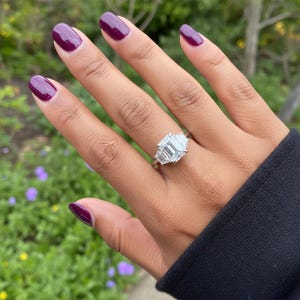 May include: A silver ring with a large, emerald-cut diamond center stone and two smaller side stones. The ring is on a hand with dark purple nail polish. The background is blurred with green foliage and purple flowers.