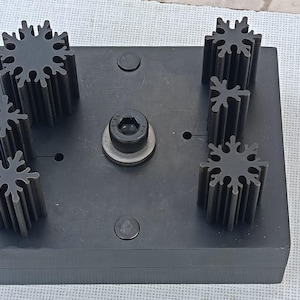 Può includere: Una base in metallo nero con diversi componenti a forma di fiocco di neve. I componenti sono disposti sulla base, che ha un bullone centrale e due elementi circolari più piccoli. I componenti sono grigio scuro.