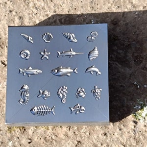 Puede incluir: Una placa de metal plateado con varios diseños de vida marina en relieve en la superficie. Los diseños incluyen peces, conchas, estrellas de mar, caballitos de mar y un cangrejo.