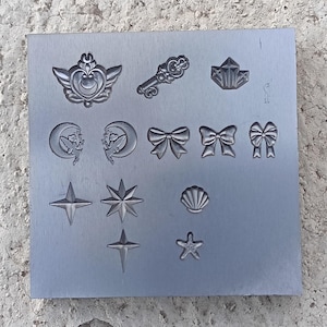 Puede incluir: Placa metálica rectangular con varios diseños en relieve, incluyendo lazos, estrellas, una llave, una concha y una luna creciente alada. La placa plateada descansa sobre una superficie clara y texturizada.