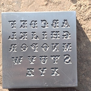 May include: A square metal stamp featuring an alphabet of decorative letters. The letters are carved into the metal, creating a negative space design. The letters are in a unique, stylized font, suitable for crafting or personalization projects.