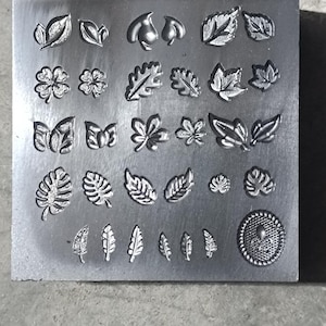 Può includere: Un timbro metallico rettangolare con vari disegni di foglie e fiori. Il timbro presenta dettagliate impressioni di diverse forme di foglie, fiori e un motivo circolare, ideale per l'artigianato o la gioielleria.