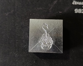 May include: A silver metal stamp with a detailed scorpion design. The stamp is square and has a smooth surface.