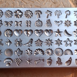 Puede incluir: Un juego de sellos de metal con una variedad de diseños, que incluyen corazones, estrellas, flores, hojas y animales. Los sellos están dispuestos en filas en una placa rectangular de metal.