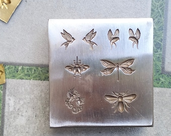 May include: A metal stamping block with six different designs: two birds, two butterflies, a dragonfly, and a bee. The designs are raised and detailed.