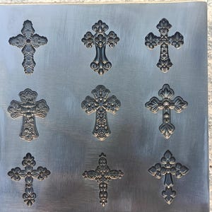 Puede incluir: Nueve cruces decorativas con diseños ornamentados dispuestas sobre una superficie de metal cepillado. Las cruces varían en estilo, con patrones florales y geométricos. El metal es de color gris plateado, y las cruces están en relieve, creando un efecto tridimensional.