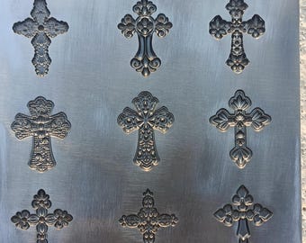 May include: Nine decorative crosses with ornate designs are arranged on a brushed metal surface. The crosses vary in style, featuring floral and geometric patterns. The metal has a silver-gray color, and the crosses are embossed, creating a three-dimensional effect.