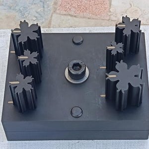 May include: A black metal tool featuring six snowflake-shaped components on a rectangular base. The base includes a central bolt and two small circular features. The snowflake components have gold-coloured accents.