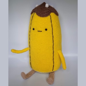May include: A crocheted banana plush toy with a brown crocheted hat and a simple face. The toy is sitting on a white surface.