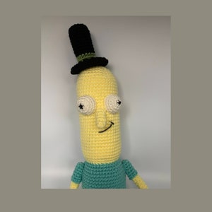 May include: A crocheted plush toy of a yellow stick figure wearing a black top hat with a green band and a teal shirt. The toy has a smiling face with white eyes and a black nose.