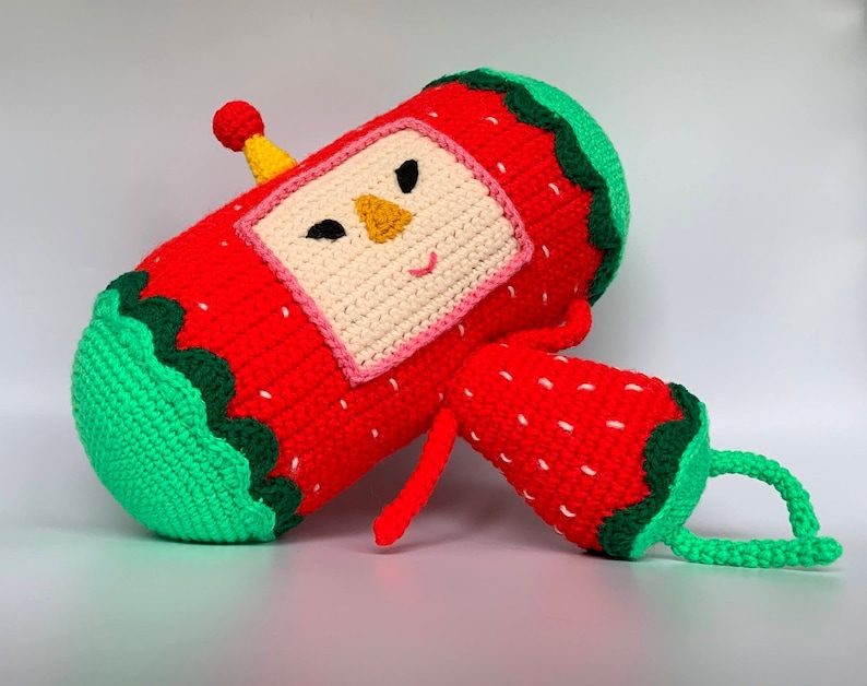 K&ouml;nnte beinhalten: Ein geh&auml;keltes Pl&uuml;schtier in Form einer Erdbeere mit gr&uuml;nem Stiel und Bl&auml;ttern. Die Erdbeere hat ein l&auml;chelndes Gesicht mit schwarzen Augen und einer gelben Nase. Das Spielzeug ist aus rotem, gr&uuml;nem, wei&szlig;em und gelbem Garn gefertigt.